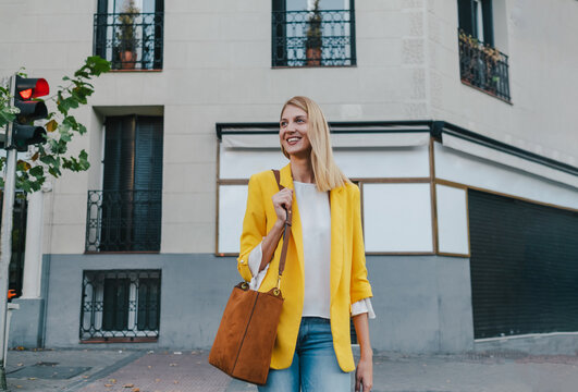 Happy young blond female in bright yellow jacket and jeans with brown handbag over shoulder smiling away while waiting to cross on traffic light on the street in the city