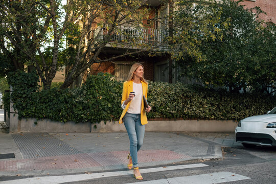 Trendy slim blond businesswoman looking away in elegant vivid yellow jacket and jeans with digital tablet walking alone along pedestrian crossing against exteriors of residential multistory buildings and cars parked on street in downtown