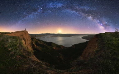 Amazing canyon with silhouette of unrecognizable person standing in cliff located near lake under colorful nigh sky with milky way on background