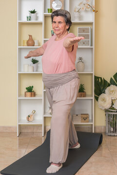 Serious senior lady in activewear standing in room near shelves with potted plants and doing stretching exercise for arms while practicing yoga at home