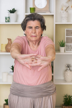 Calm elderly lady standing near shelves with potted plants and doing stretching exercise for arms while practicing yoga at home