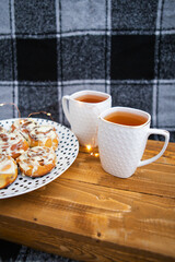 Two cups of black tea stand on a wooden tray on the sofa with a black and white checkered plaid. Fresh and fragrant cinnamon rolls close-up lie on a plate with polka dots. The garland is blinking