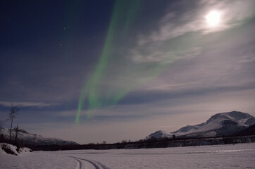 aurora borealis, northern light over a frozen snowy river bed with full moon and majestic mountain