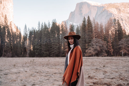 Happy relaxed young female traveler in stylish outfit sitting on stone border against picturesque mountain scenery with rocky cliffs and coniferous forest in Yosemite National Park in USA