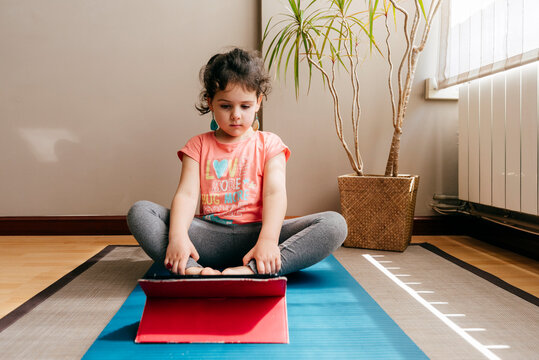 Thoughtful little girl sitting on mat near window at home resting after practicing yoga throughout video tutorials on tablet