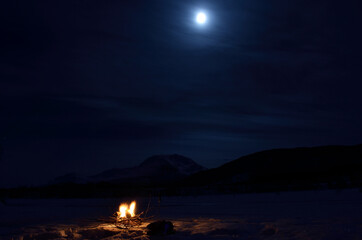 burning fire on frozen river bed with majestic snowy mountain and full moon in the background