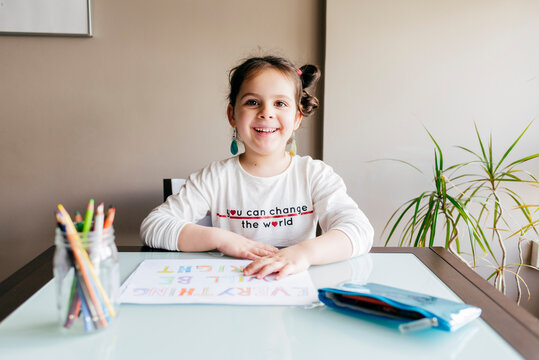 little girl in casual clothes sitting at wooden table and drawing picture with colored pencils while spending time at home