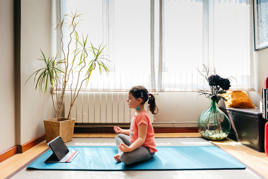 Little girl sitting in lotus pose and meditating on mat with eyes closed at home with big window while practicing yoga and watching video tutorials on tablet