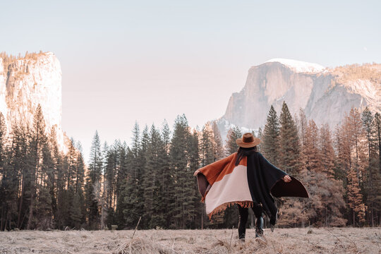 Happy relaxed young female traveler in stylish outfit sitting on stone border against picturesque mountain scenery with rocky cliffs and coniferous forest in Yosemite National Park in USA