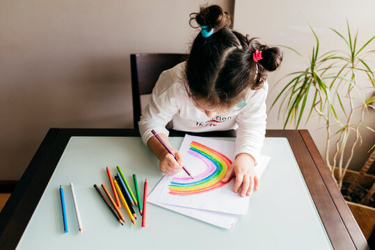 From above of anonymous girl in casual clothes  sitting on chair at wooden table and drawing colorful rainbow with pencil on paper
