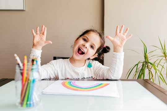 Playful little girl in casual clothes showing hands and laughing while sitting at wooden table and drawing rainbow with pencils at home