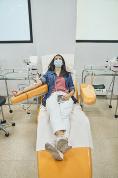 From Above Of Relaxed Young Female In Protective Mask With Smartphone Sitting In Medical Chair During Blood Donation In Modern Hospital