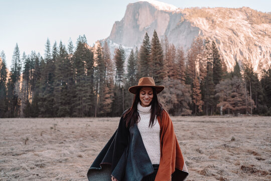 Happy relaxed young female traveler in stylish outfit sitting on stone border against picturesque mountain scenery with rocky cliffs and coniferous forest in Yosemite National Park in USA