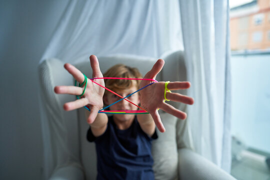 Closeup of hands of boy playing cats cradle game and demonstrating basic figure made with colorful string while spending free time at home looking at camera