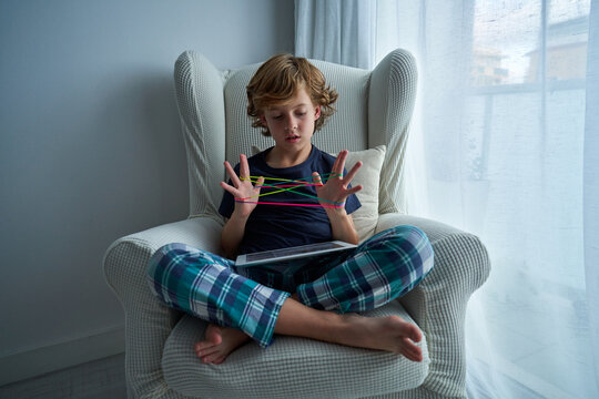 Focused school aged boy in pajamas sitting on armchair and watching video on tablet while playing cats cradle game and creating figures with string