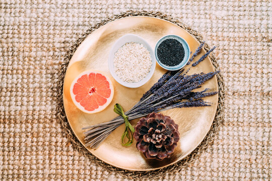 From above of gold plate with half of freshly juicy grapefruit with fragrant spruce cone next to small bouquet of dried lavender and two ceramic bowls with chia and sesame seeds locating on wicker rattan stand on rug