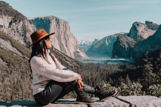 Full body side view of happy relaxed young female traveler in stylish outfit sitting on stone border against picturesque mountain scenery with rocky cliffs and coniferous forest in Yosemite National Park in USA