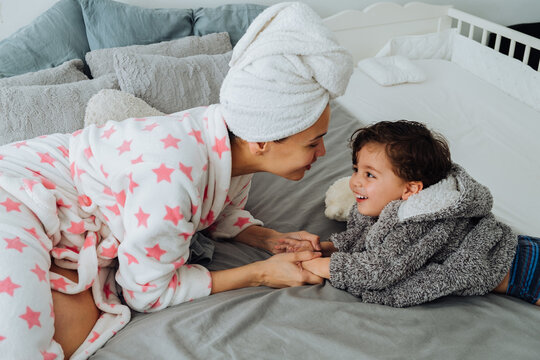 Cheerful woman in bathrobe having fun with little boy on soft bed looking at each other