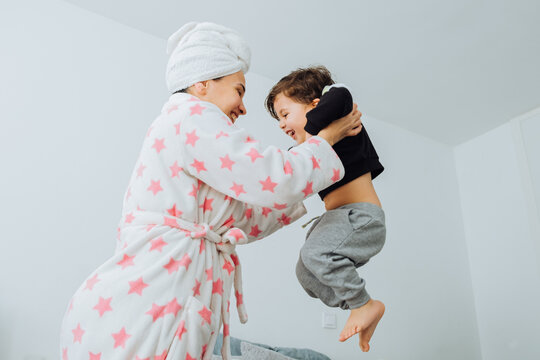 From below side view of cheerful woman in bathrobe tossing little son while playing together during weekend at home looking at each other