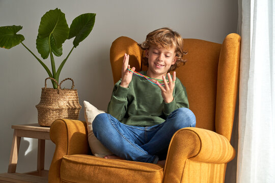 Cheerful preteen boy in casual clothes sitting in cozy armchair and playing interesting cats cradle game while resting at home