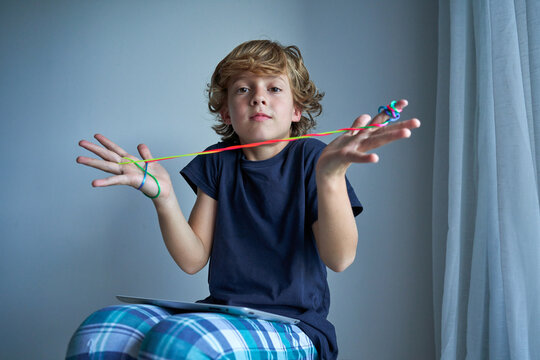 Excited schoolboy in pajamas with tablet on knees creating difficult figure with string while playing cats cradle game at home sitting on stool