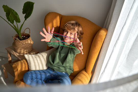 From above cheerful preteen boy in casual clothes sitting in cozy armchair and playing interesting cats cradle game while resting at home looking at camera