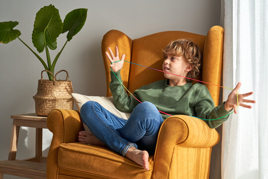 Cheerful preteen boy in casual clothes sitting in cozy armchair and playing interesting cats cradle game while resting at home looking away
