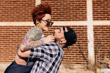 Side view of happy young man embracing hipster tattooed girlfriend while having fun during romantic date on city street near brick building