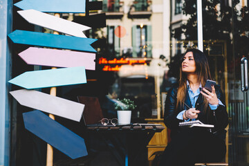 Confident young woman blogger with digital smartphone device looking away while working remotely in cafe outdoors and using free high speed 4G internet connection.Copy space area for advertising