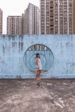 Female in summer dress standing on concrete base of unusual city installation in shape of passage with round holes and looking at camera