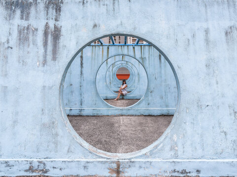 Female in summer dress sitting on concrete base of unusual city installation in shape of passage with round holes and looking away