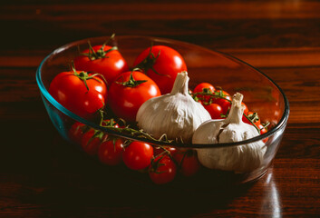 Un recipiente de cristal con tomates, tomats cherry y ajos sobre una mesa de madera marr&oacute;n oscuro.