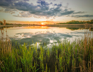 Scenic view at beautiful spring sunset with reflection on a shiny lake with green reeds, bushes, grass, golden sun rays, calm water ,deep blue cloudy sky , glow on background, spring evening landscape