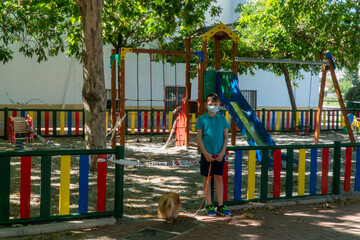 Fototapeta na wymiar A sad child is standing at the quarantined children's playground. COVID-19 security and protection measures in the city park.
