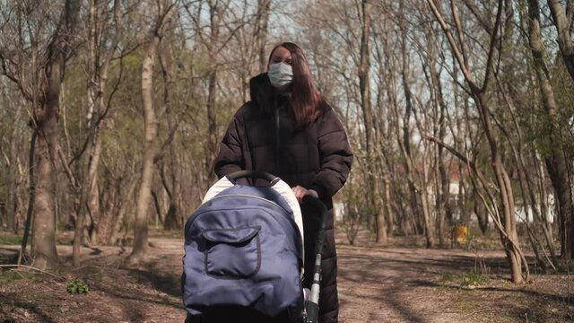 Mom in a protective mask with a pram is walking in the park among the trees. Dramatic Shot, Pandemic Virus, 4K.