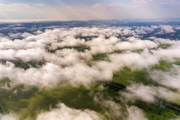 Aerial view of white clouds above a town or village with rows of buildings and curvy streets between green fields in summer. Countryside landscape from above.