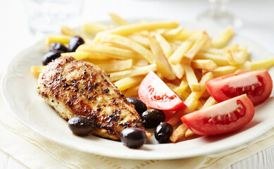 Fried chicken breast with french fries, tomatoes and olives. White wooden background. Close up.