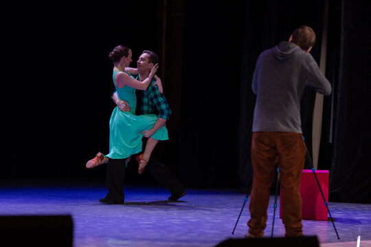 A pair of dancers a man and a woman are dancing on stage and a man videographer records a video clip and reports.