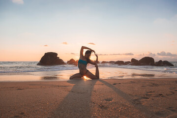 Fototapeta premium Young attractive latin woman wearing a bikini practicing yoga on the beach at sunset, healthy mental and body lifestyle concept. Yogi pose