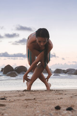 Young attractive latin woman wearing a bikini practicing yoga on the beach at sunset, healthy mental and body lifestyle concept.