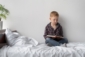 Schoolchild at home in quarantine. Distance learning. A blond boy is sitting on the bed and reading a book. The child stays at home and does the homework himself. 