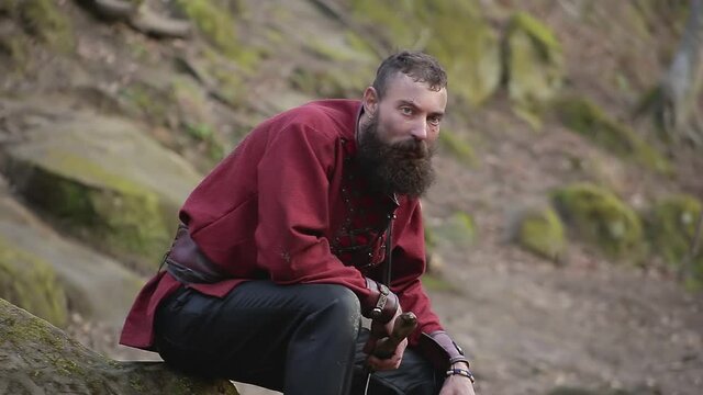 Brutal man with big beard in clothes in the image of Viking with axe in his hands sits on stone and breathes heavily in the forest
