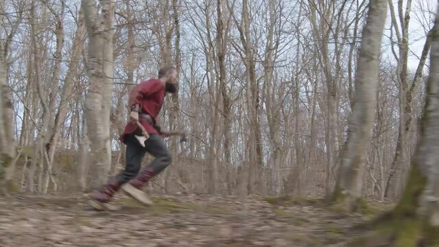 Slow motion brutal man with long beard in suit and the image of Viking with axes in his hands makes his way through thicket and runs through forest