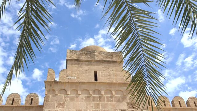 Ancient walls of antique fortrees isolated at sunny blue sky background. Green foliage of palm trees in foreground. Tunisia.