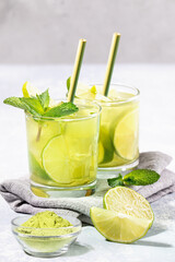 Two glasses of iced green matcha tea with lime, ice, mint, matcha powder, bamboo drinking straws on napkin on light grey backdrop. Healthy food, No Plastic, Zero Waste. Vertical shot. Selective focus.
