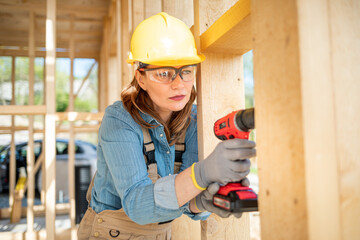 Female worker using cordless drill on construction site of wood frame house