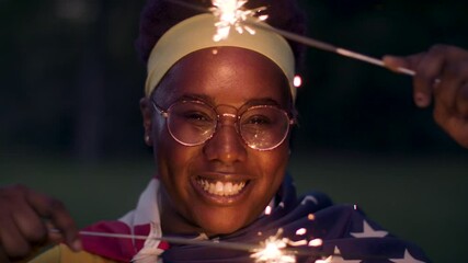 Celebrating Fourth of July. African American woman smiles draped in the flag-waving sparklers. Shot in slow-motion and in 4k. - Powered by Adobe