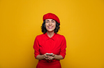 Such a nice trip. Half-length photo of a cute smiley french style dressed woman in a red beret, holding her phone and looking straight.