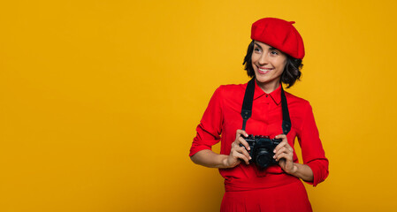 Ready to make a photo. Half-length photo of a smiley travelling woman french style dressed, carrying a camera on a strap on her neck, looking aside with shiny eyes.