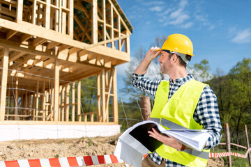 Engineer with hardhat and blueprints on building site of wood frame house under construction